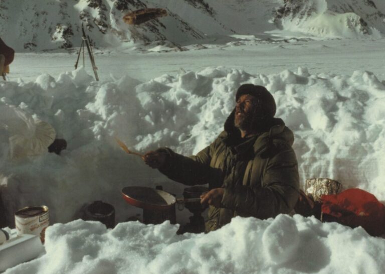 穿绿羽绒服的男子在雪地营地挥铲做饭